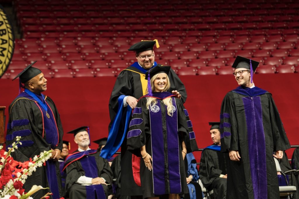 Alabama Law student smiles as she is hooded at commencement. Professors standing on the stage watch smiling at the graduate.
