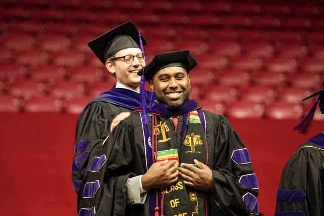 Alabama Law student smiles with professor standing behind him after being hooded at commencement ceremony.