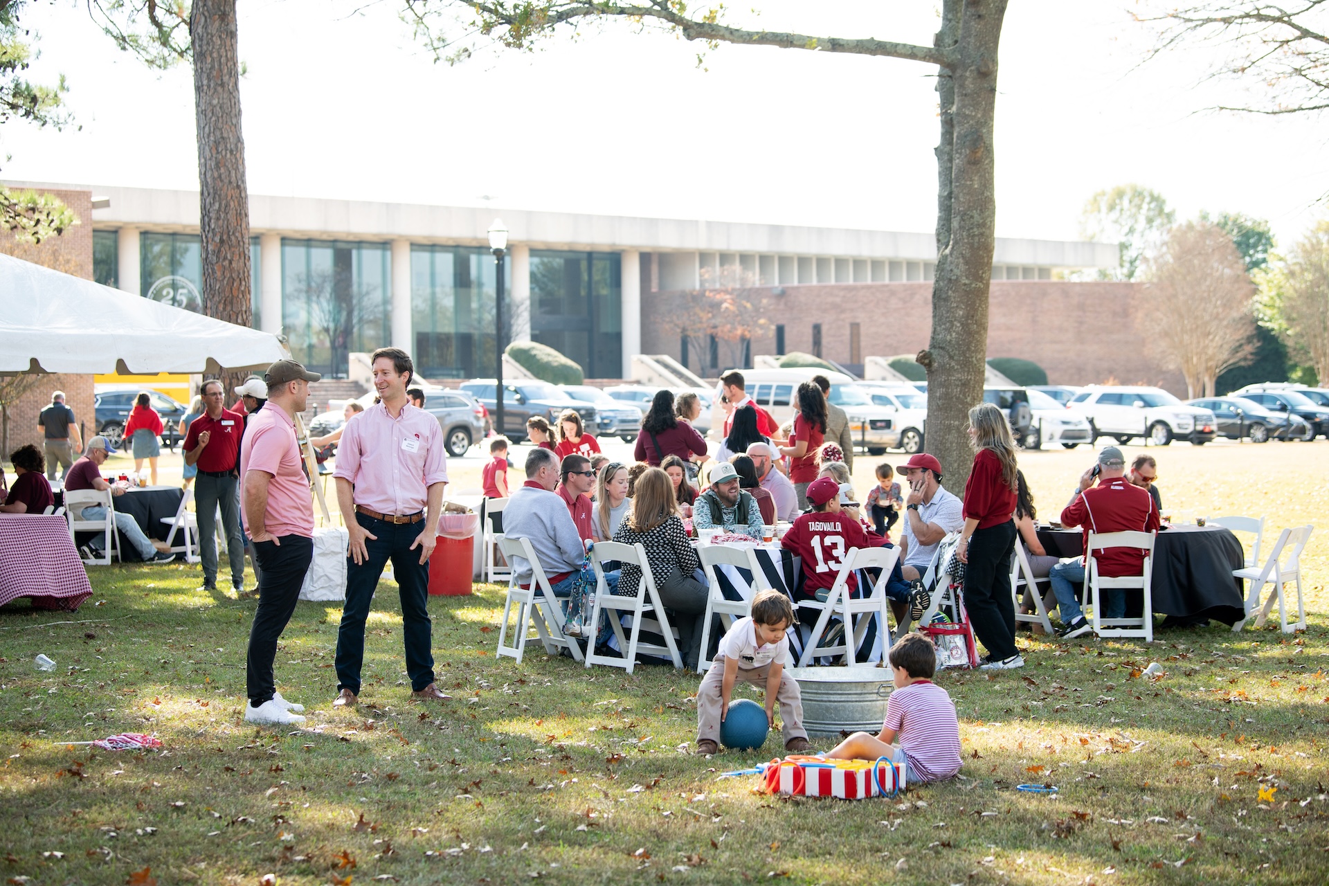 a tailgate on the front law of the University of Alabama School of Law with the Law School building visible in the background. There are people sitting at tables and children playing with toys on the lawn.