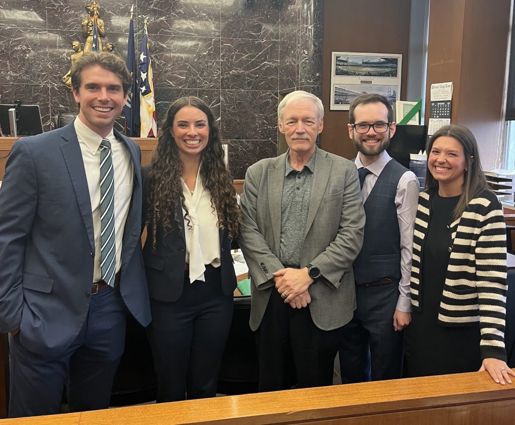 A group of five people stand at the front of a courtroom in business professional attire
