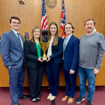 a group of five people pose in a courtroom. the two women in the middle are holding a trophy.