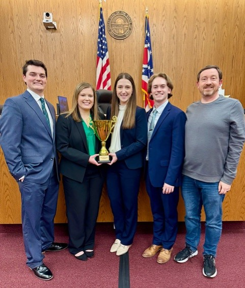 a group of five people stands in a row. two women in the middle are holding a trophy. 