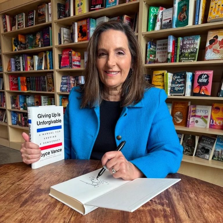 Joyce Vance holds a copy of her book, Giving Up Is Unforgivable, while signing another copy. She is in front of a bookshelf with many other books displayed. 
