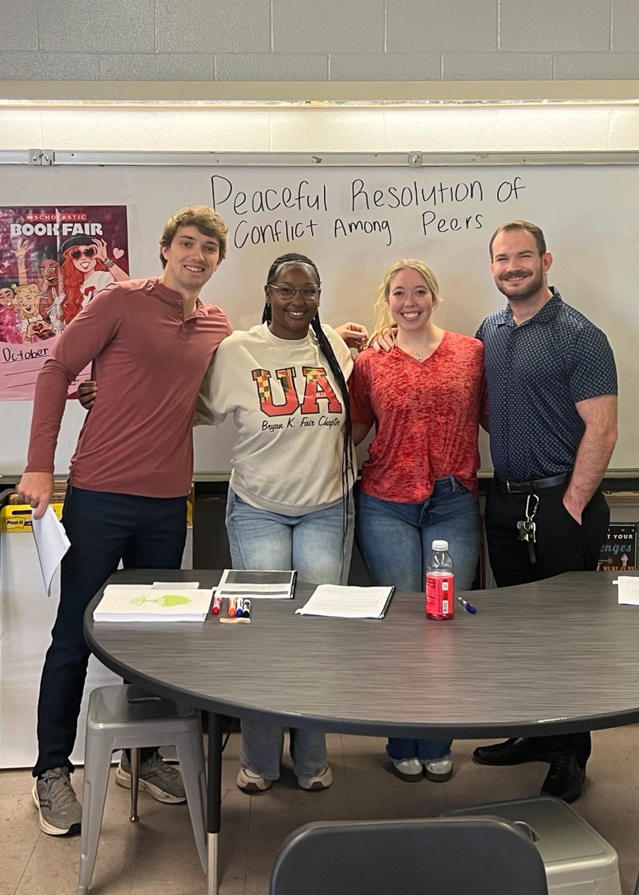 Four young adults stand at the front of a classroom. Behind them, 