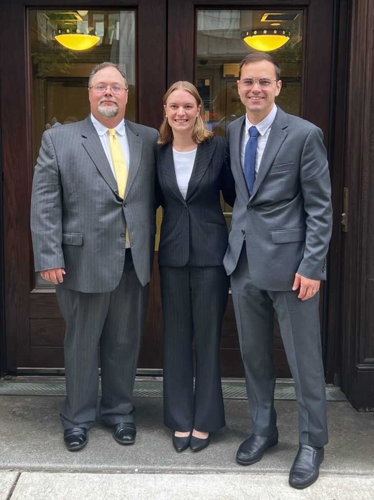Three people dressed in suits stand in front of a door with 