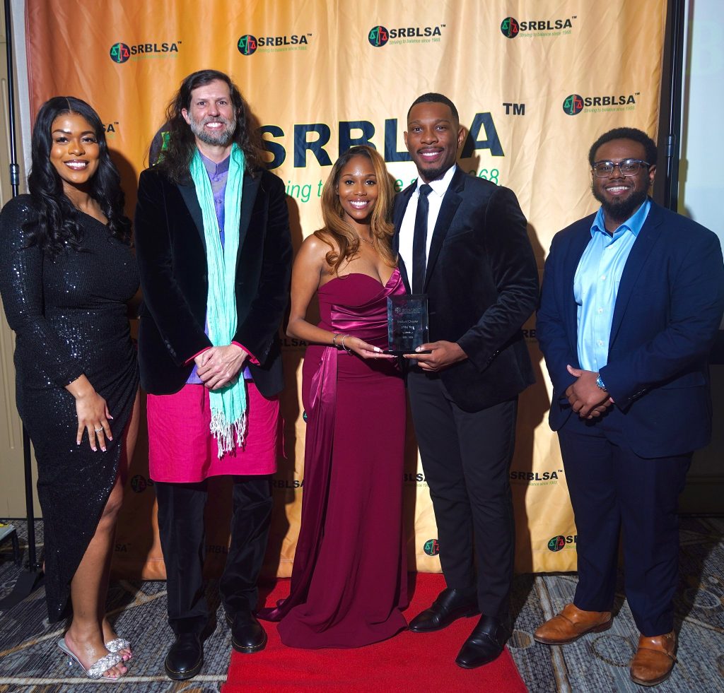 Five people standing in a line in formal dress. They are in front of a backdrop with the SRBLSA logo printed on it. The two people in the middle are holding an award. 