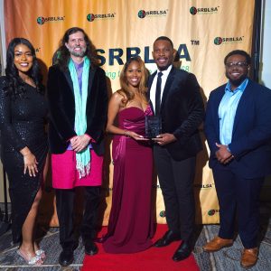 Five people standing in a line in formal dress. They are in front of a backdrop with the SRBLSA logo printed on it. The two people in the middle are holding an award.