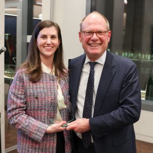A young woman wearing a pink tweed jacket poses with a man dressed in a suit and tie. They are holding a glass award and smiling at the camera.