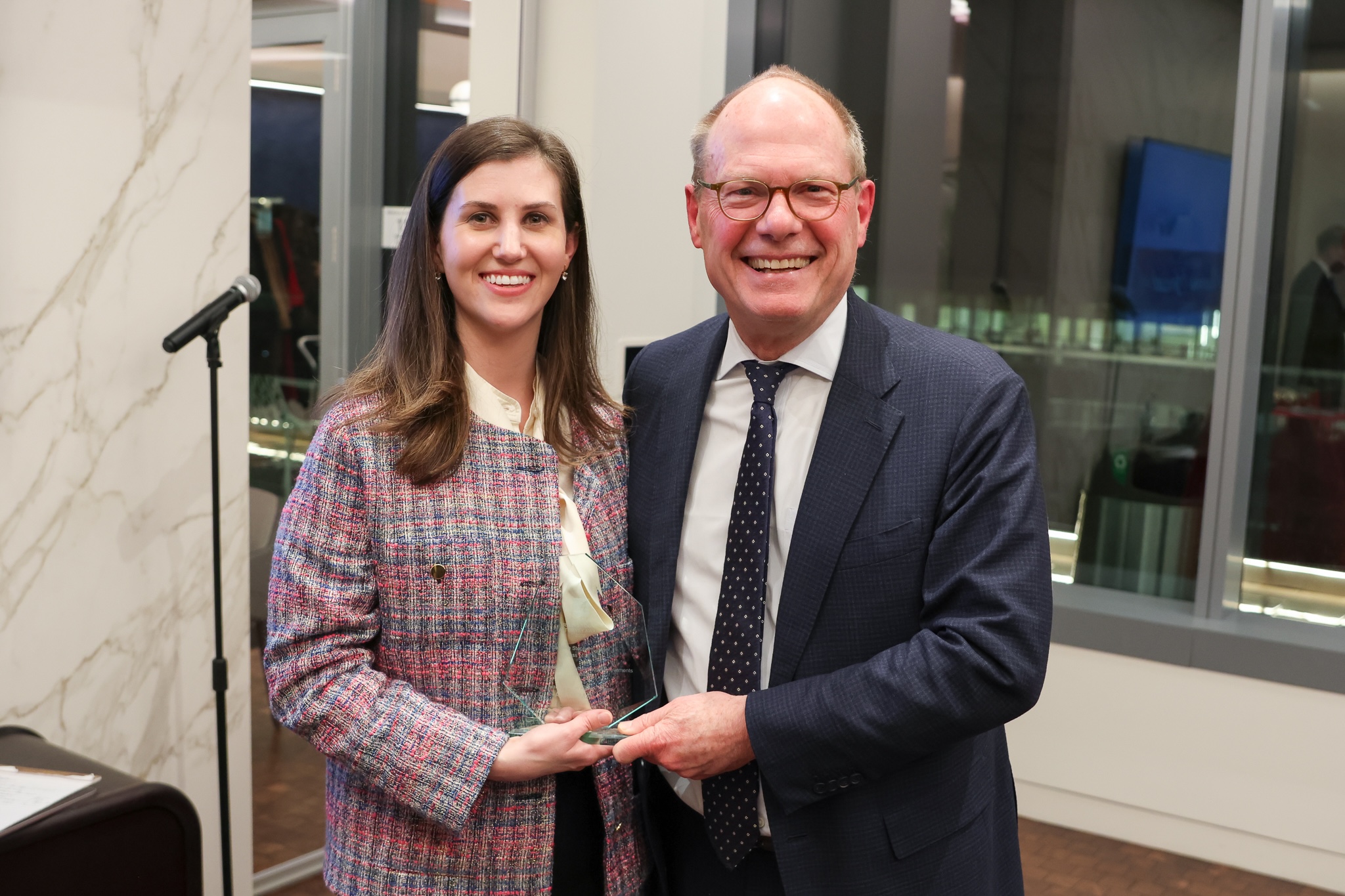 A young woman wearing a pink tweed jacket poses with a man dressed in a suit and tie. They are holding a glass award and smiling at the camera. 